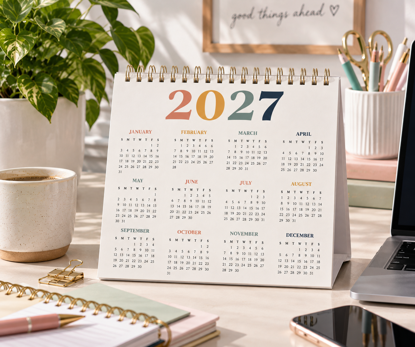 A colorful 2027 desk calendar on a sunny desk with coffee, a plant and notebooks